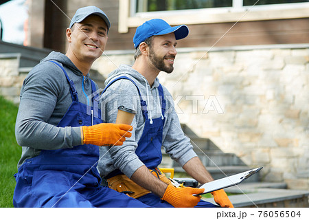Responsible contractors. Happy young workmen in uniform taking a break, sitting outdoors having coffee and holding papers while working on construction project 76056504
