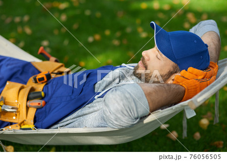 Peaceful young male builder wearing uniform taking a break, lying in a hammock outdoors with eyes closed on a sunny day 76056505