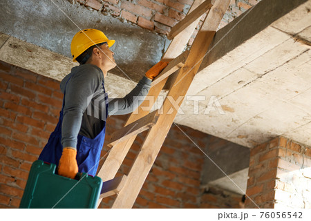 From the ground up. Young male builder in blue overalls and hard hat climbing up the ladder, holding toolbox while working at construction site From the ground up. Young male builder in blue overalls and hard hat climbing up the ladder, holding toolbox while working at construction site 76056542