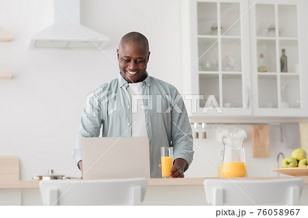 Happy african american mature man drinking orange juice and watching movie on laptop in kitchen 76058967