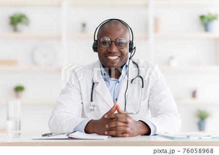 Family doctor consulting remotely. Smiling african american pediatrician in headphones sitting at table in living room 76058968