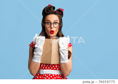 Surprised pin up woman in polka dot dress and glasses holding book over blue studio background, copy space 76059775