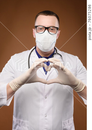 A young doctor in a protective mask and a white coat and glasses with a stethoscope around his neck shows the heart symbol and shape with his hands. 76075286