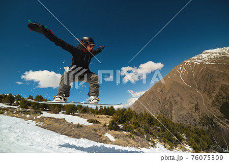 Snowboarder woman jumping from a kicker springboard from the snow on a sunny day in the mountains in a homemade snowboard park 76075309