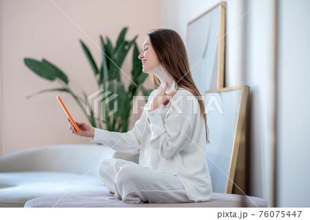 Young woman in white sitting on a soft chair and making a photo of tapes on her neck Young woman in white sitting on a soft chair and making a photo of tapes on her neck 76075447