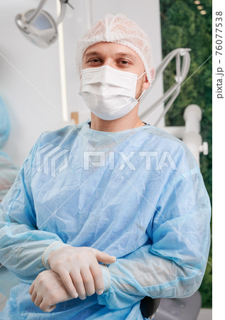 Portrait of young male dentist wearing disposable clothes sitting in modern clinic 76077538