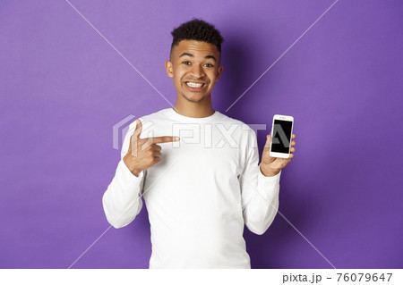 Portrait of handsome african-american man, smiling and pointing finger at smartphone screen, showing application, standing over purple background Portrait of handsome african-american man, smiling and pointing finger at smartphone screen, showing application, standing over purple background 76079647
