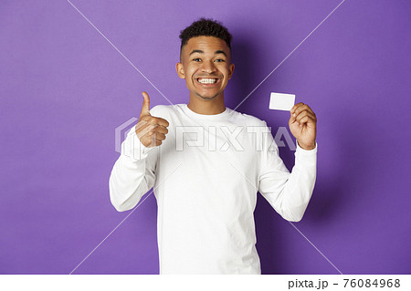 Portrait of amazed african-american man showing thumbs-up and credit card, recommend bank loan, standing over purple background Portrait of amazed african-american man showing thumbs-up and credit card, recommend bank loan, standing over purple background 76084968