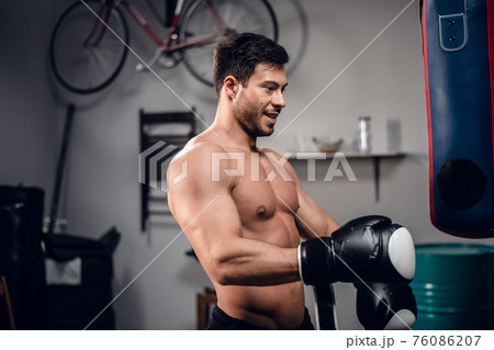a young athlete with a muscular torso is warming up with a punching bag in his garage. a young athlete with a muscular torso is warming up with a punching bag in his garage. 76086207