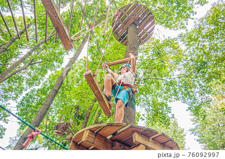 a young boy in outfit and a helmet holds on to the ropes near the round wooden platform in Forest adventure park 76092997