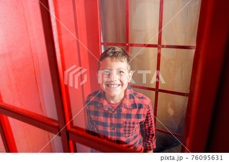 a cheerful smiling fair-haired teenage boy, dressed in a red and black plaid shirt, stands in a red telephone box 76095631