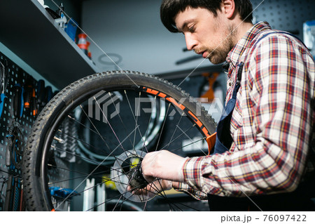 Bicycle mechanic fixing cycle wheel in repair shop. Velocipede service. Repairer male bike shop engineer fix bicycle wheel in workshop. Technician fixes bike parts. Handyman at work in the workplace Bicycle mechanic fixing cycle wheel in repair shop. Velocipede service. Repairer male bike shop engineer fix bicycle wheel in workshop. Technician fixes bike parts. Handyman at work in the workplace 76097422