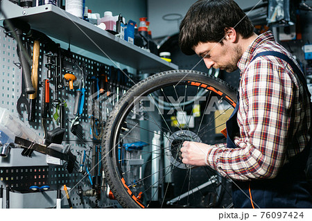 Male mechanic working in bicycle repair shop, mechanic repairing bike using special tool, wearing protective gloves. Young attractive serviceman fixing customer's bicycle wheel at his own workshop Male mechanic working in bicycle repair shop, mechanic repairing bike using special tool, wearing protective gloves. Young attractive serviceman fixing customer's bicycle wheel at his own workshop 76097424