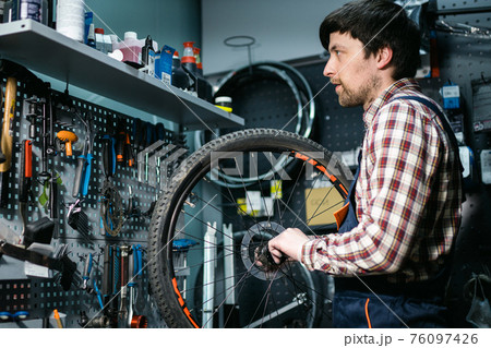 Young caucasian repairman repairing mountain bike wheel in bike shop workshop. Fixing bicycle. Caring for you wheels. man mechanic working in garage. Worker fixing cycle gear. Small business theme Young caucasian repairman repairing mountain bike wheel in bike shop workshop. Fixing bicycle. Caring for you wheels. man mechanic working in garage. Worker fixing cycle gear. Small business theme 76097426