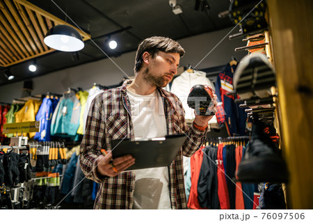 Sporting goods store owner with clipboard checking inventory. Tourist store manager working near showcase with hiking boots makes check list of orders. Salesman exhibit display of walking boots 76097506
