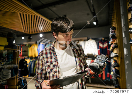 Sales assistant sports travel store makes inventory hiking boots. Travel goods store theme. Clothes and footwear for sports and travel. Store manager near shelves with hiking boots makes check list 76097977