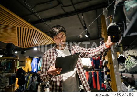 Sales assistant sports travel store makes inventory hiking boots. Travel goods store theme. Clothes and footwear for sports and travel. Store manager near shelves with hiking boots makes check list Sales assistant sports travel store makes inventory hiking boots. Travel goods store theme. Clothes and footwear for sports and travel. Store manager near shelves with hiking boots makes check list 76097980
