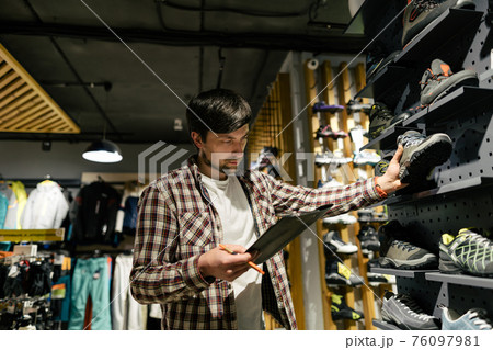 Business owner in sports store works with showcase trekking and mountaineering boots for sale, holding clipboard in his hands, checks availability of goods and collects to process online orders Business owner in sports store works with showcase trekking and mountaineering boots for sale, holding clipboard in his hands, checks availability of goods and collects to process online orders 76097981