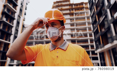 Topic is health protection in production and construction. Caucasian male worker in orange uniform and protective respirator kn 95 puts on construction orange hard hat and poses at construction site Topic is health protection in production and construction. Caucasian male worker in orange uniform and protective respirator kn 95 puts on construction orange hard hat and poses at construction site 76098019