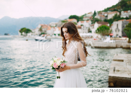 The bride holds a bouquet of roses in her hands and stands on the pier near the old town of Perast  76102021