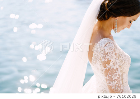 Close-up of the neck and neck of the bride, face profile. Beautiful big earring in ears, shoulder with white lace dress on background of glare of water. Fine-art wedding photo in Montenegro, Perast. Close-up of the neck and neck of the bride, face profile. Beautiful big earring in ears, shoulder with white lace dress on background of glare of water. Fine-art wedding photo in Montenegro, Perast. 76102029