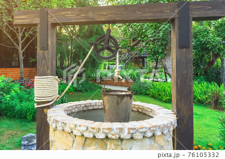 Beautiful artesian well made by stones and wheel pulley with metal bucket and rope in peaceful garden atmosphere. Retro stone water well in rural area. Garden decoration with antique items. 76105332