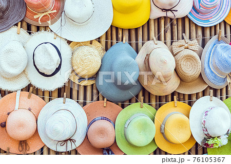 A variety of fashionable and colorful hats hanging on the bamboo backdrop of curbside shop.Women's designer hats from the sun of different colors. Design of women's beach hats. Beach hats for summer. 76105367