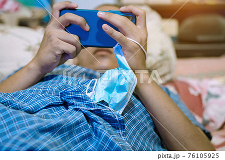 A surgical mask to prevent infection of Coronavirus(C0VID-19) hung on hand of young man while lying on the bed to play game with mobile phones. New normal lifestyle. Selective focus on surgical mask. A surgical mask to prevent infection of Coronavirus(C0VID-19) hung on hand of young man while lying on the bed to play game with mobile phones. New normal lifestyle. Selective focus on surgical mask. 76105925