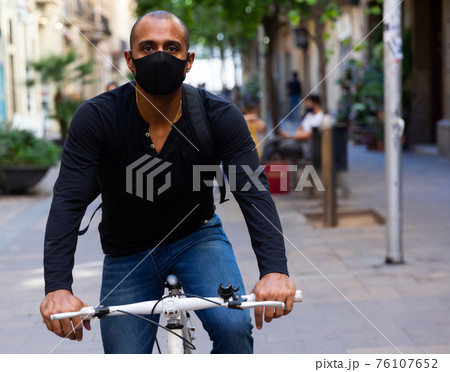 Latin american man in protective mask riding bike on city street Latin american man in protective mask riding bike on city street 76107652