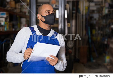 Warehouse male worker in protective mask and overalls counts the quantity of goods and writes in notebook Warehouse male worker in protective mask and overalls counts the quantity of goods and writes in notebook 76108069