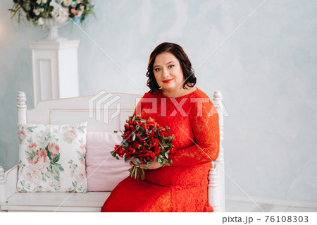 a woman in a red dress sits on a sofa and holds a bouquet of red roses and strawberries in the interior. a woman in a red dress sits on a sofa and holds a bouquet of red roses and strawberries in the interior. 76108303