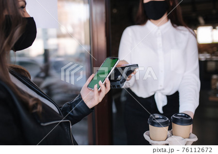 Woman standing outdoors by cafe and paying with smartphone during Covid-19 pandemic. Cashier hand holding credit card reader machine while client holding phone for NFC payment. Green screen on phone 76112672