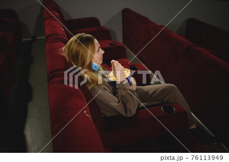 young woman in medical mask eating popcorn sitting in the cinema young woman in medical mask eating popcorn sitting in the cinema 76113949