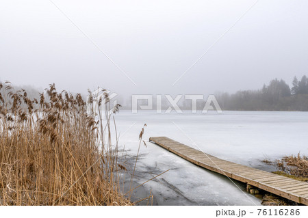 Wooden footbridge on lake with thick mist foggy air over frozen water 76116286