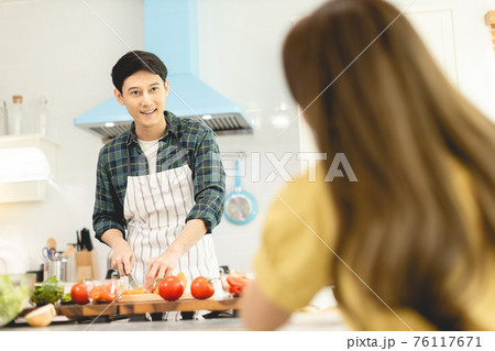 Husband slicing vegetables to prepare breakfast. With a young wife supporting him In a romantic atmosphere Husband slicing vegetables to prepare breakfast. With a young wife supporting him In a romantic atmosphere 76117671