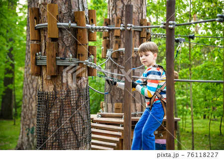 Young cute child boy in summer clothing, safety harness and helmet attached with carbine to cable moves slowly along rope way on green trees sunny bokeh background. Sport, game, leisure concept 76118227