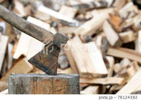 Old rusty axe stuck in a log with a pile of chopped firewood on the background 76124392
