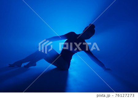 Ballerina in black bodysuit dancing with blue light and smoke. Silhouette of dancer in ballet shoes. 76127273