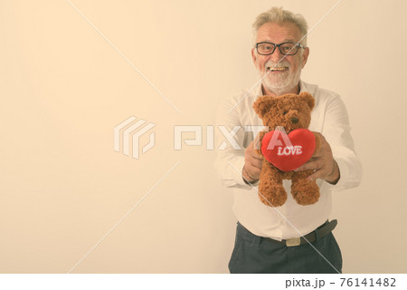 Studio shot of happy senior bearded man smiling while giving teddy bear with heart and love sign 76141482