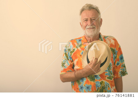 Studio shot of handsome senior bearded tourist man holding hat on his chest against white background 76141881
