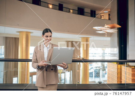 Young businesswoman in beige jacket standing with a laptop 76147957