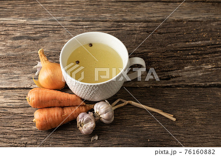 Bowl with clear rich broth. White crockery on an aged wooden table. Vegetables - carrots, onions, garlic, spices 76160882