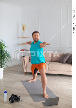 Middle aged man works out at home with an online tutorial. He performs a balance exercise in front of a laptop monitor. Vertical photo. 76161369