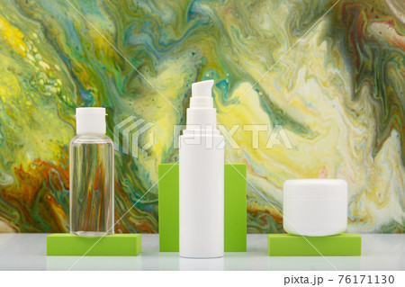 Skin lotion in transparent bottle, face cream and lip balm on green podiums against marbled background in green colors.  76171130