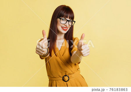 Young caucasian businesswoman in trendy yellow dress and eye glasses, isolated on yellow background, giving a thumbs up gesture and smiling with teeth to camera Young caucasian businesswoman in trendy yellow dress and eye glasses, isolated on yellow background, giving a thumbs up gesture and smiling with teeth to camera 76183863
