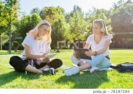 Girls teenage students sitting on green lawn in park with backpack, digital tablet 76187284