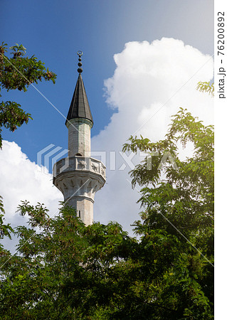 minaret protrudes from the trees, against a blue sky with clouds, sun glare minaret protrudes from the trees, against a blue sky with clouds, sun glare 76200892