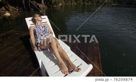 Woman lie on a sunbed in sunglasses and a boho silk shawl. Girl rest on a flood wood underwater pier. The pavement is covered with water in the lake. 76209874