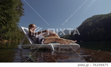 Woman lie on a sunbed in sunglasses and a boho silk shawl. Girl rest on a flood wood underwater pier. The pavement is covered with water in the lake. Woman lie on a sunbed in sunglasses and a boho silk shawl. Girl rest on a flood wood underwater pier. The pavement is covered with water in the lake. 76209875