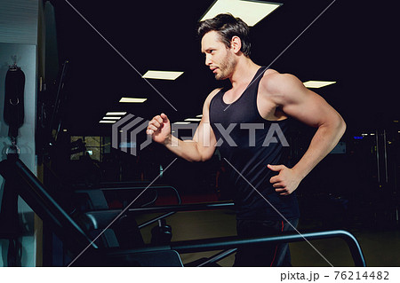 Man athlete runs jogging on a treadmill in dark gym Man athlete runs jogging on a treadmill in dark gym 76214482